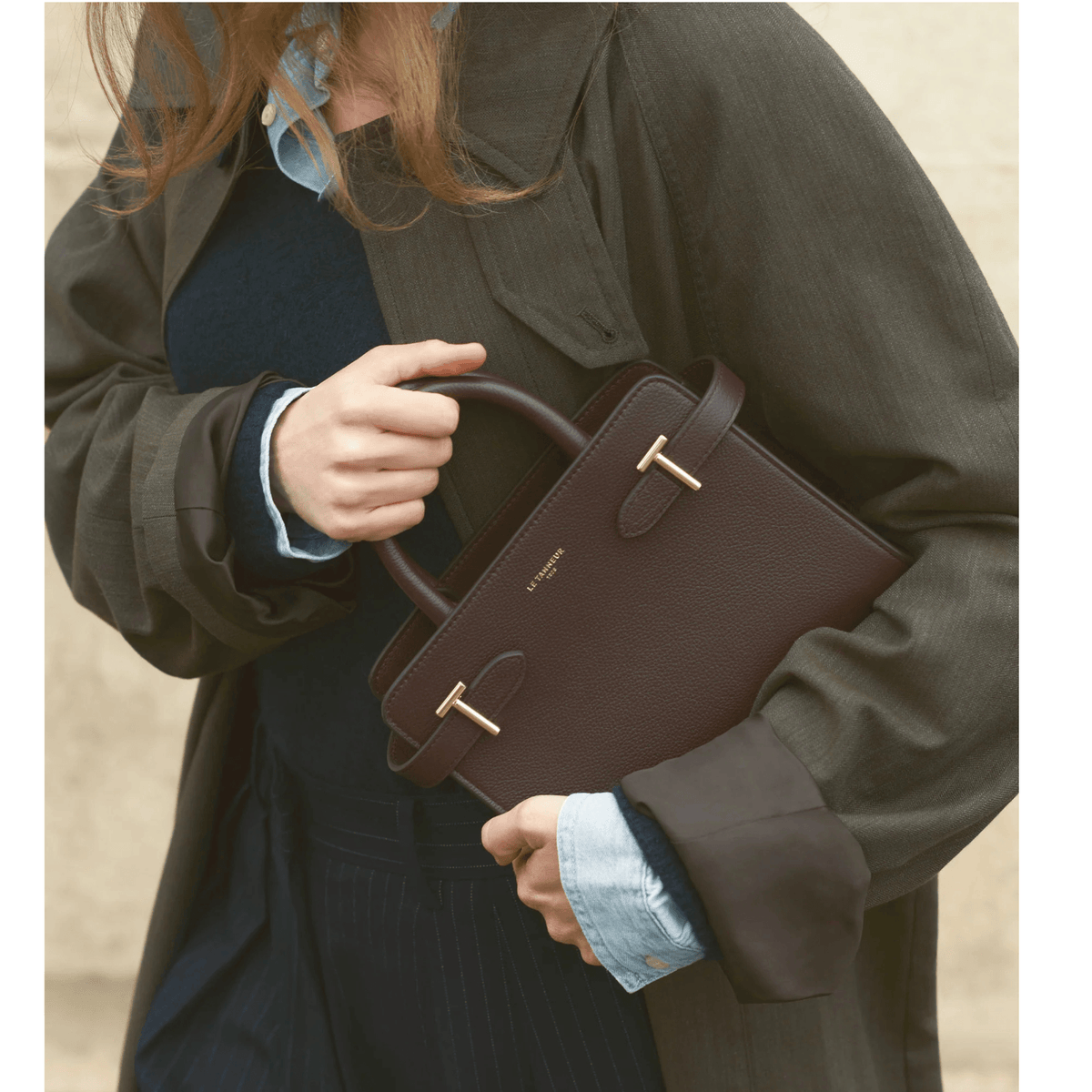 Femme portant à la main le petit sac Émilie Le Tanneur en cuir grainé marron foncé avec détails dorés